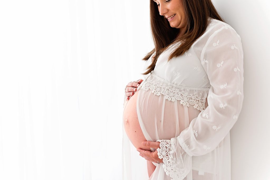 A pregnant lady at her Maternity photo shoot in a white lacy dress holding her baby bump