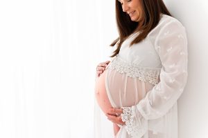 A pregnant lady at her Maternity photo shoot in a white lacy dress holding her baby bump